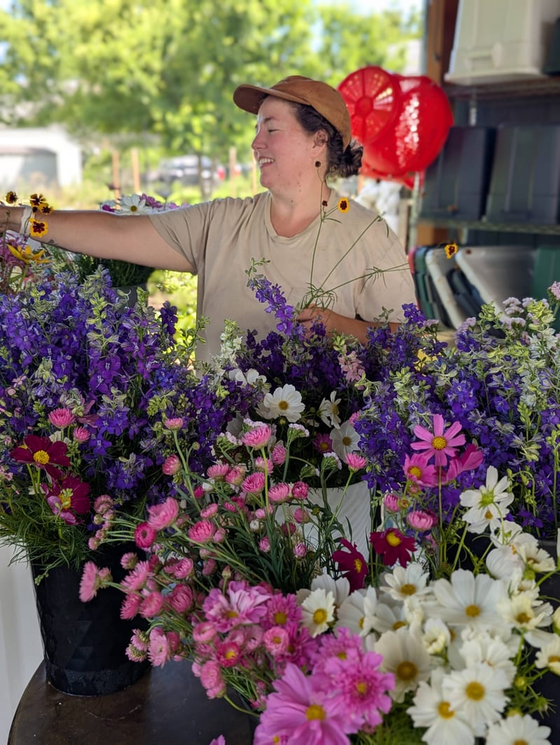 Emma reaching across buckets of delphinium, cosmos and strawflower at the farm stand